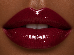 Deep complexion lips wearing JJ Luxe Cosmetics Nocturne Sheer Peptide Glaze in a glossy deep burgundy wine-plum shade.